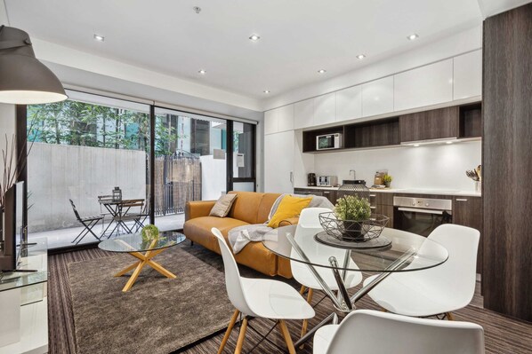 Une cuisine moderne avec des meubles blancs, un coin repas doté d’une table en verre et de chaises blanches, et un canapé agrémenté d’un coussin jaune.