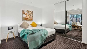 Une chambre d’hôtel moderne, équipée d’un lit, d’une table de chevet, d’une lampe et d’un grand miroir qui reflète la pièce.