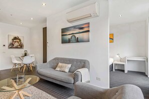 A modern living room with a grey sofa, a glass coffee table, and a painting of a sunset on the wall.