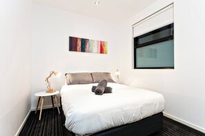 A modern hotel room with a large bed, a small bedside table, a lamp, a window, and a colorful abstract painting on the wall.