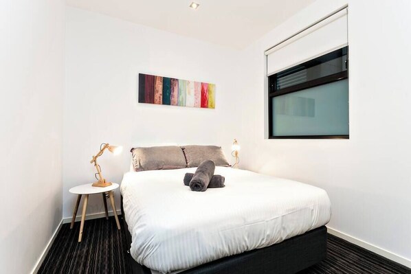 A modern hotel room with a large bed, a small bedside table, a lamp, a window, and a colorful abstract painting on the wall.