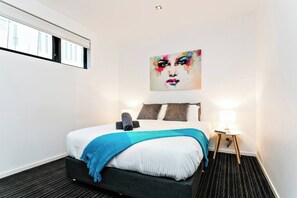 Una camera d'albergo con un letto, un quadro appeso al muro, una finestra e un comodino con una lampada.