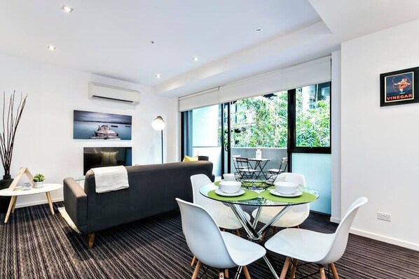 A modern room with a sofa, a dining table with chairs, a TV, and a large window with a view of greenery.