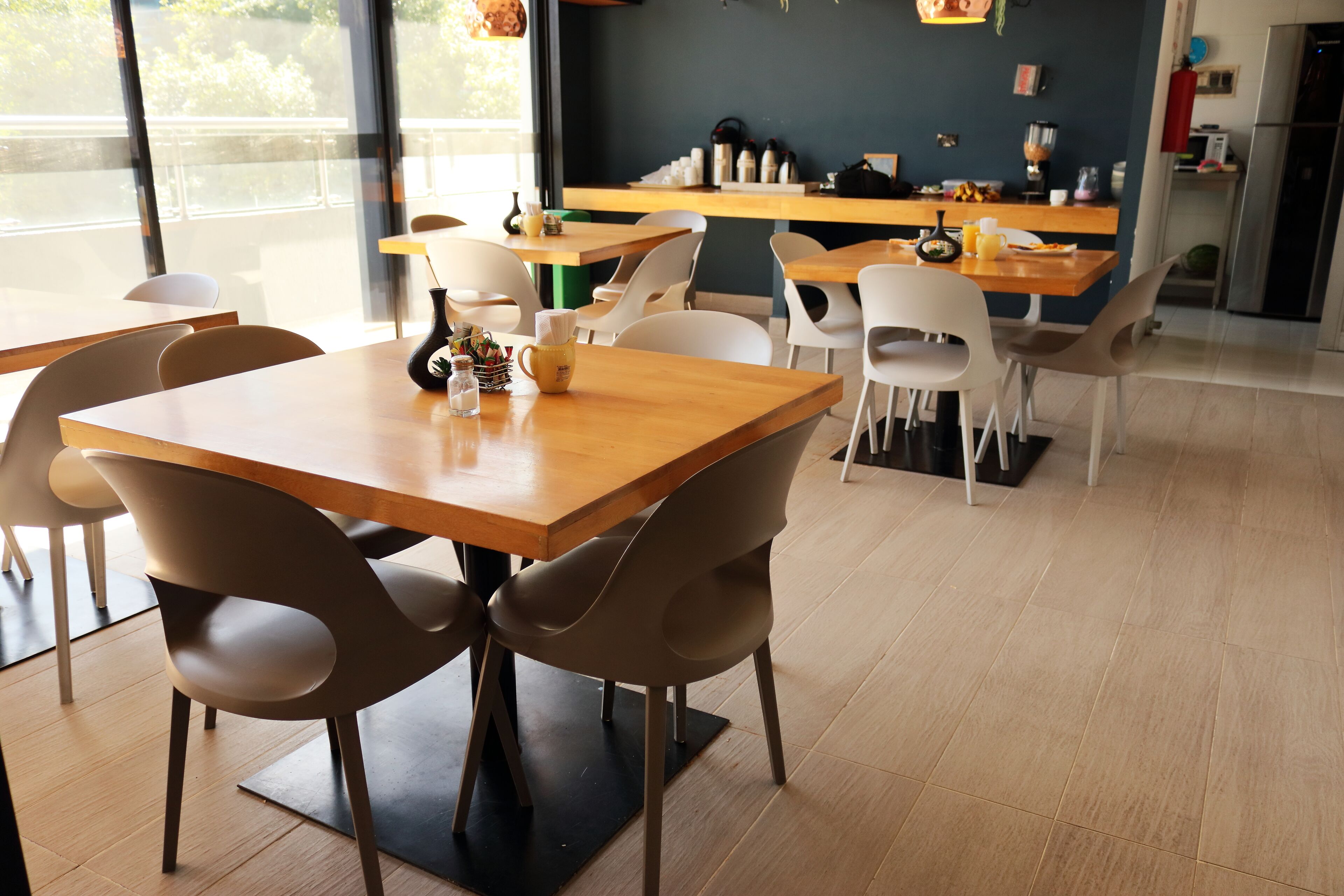 A modern cafe with wooden tables and chairs, a kitchen area in the background, and large windows providing natural light.