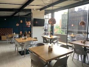 A modern cafe with wooden tables and chairs, a large window with a city view, and a black metal shelf with decorative items. - Hotel Loup (Medellín)