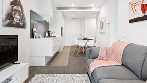 A modern living room with a grey sofa, a flat-screen TV, and a kitchen area with white cabinets and a dining table. - Sierra - Resort Living * Gym Sauna Pool WiFi (Southbank)