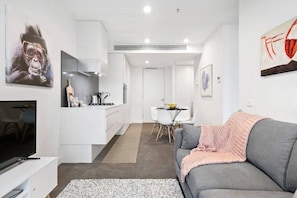 Un salon moderne avec un canapé gris, un téléviseur à écran plat et un coin cuisine doté de meubles blancs et d’une table à manger.