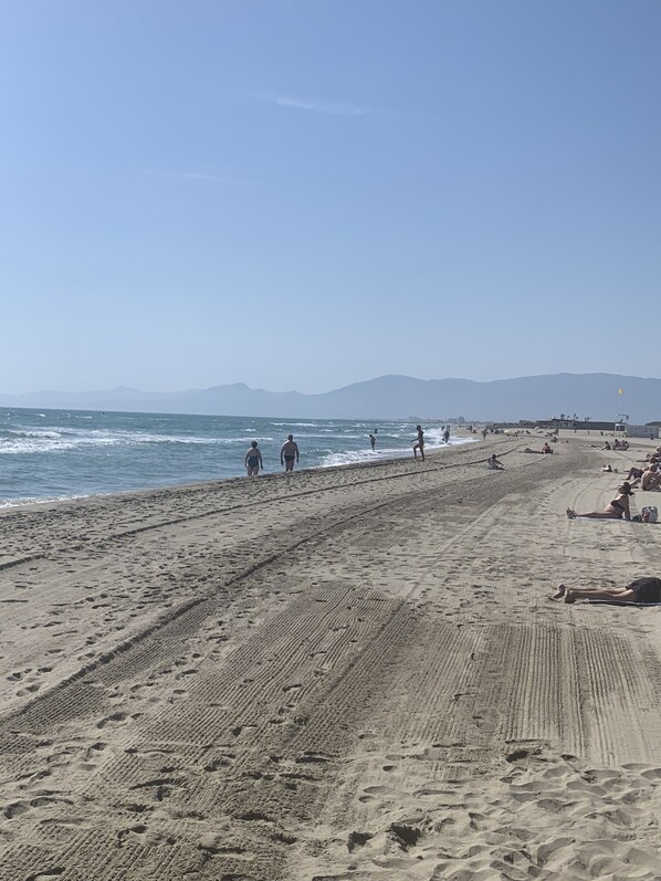 A wide sandy beach with people walking and sunbathing, a clear sky, and distant mountains.