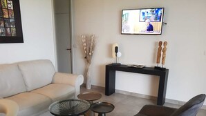 Een moderne woonkamer met een beige bank, een zwarte fauteuil, een glazen salontafel en een klein rond tafeltje. Tegen de muur staat een zwarte dressoir met twee houten sculpturen, en erboven is een flatscreen televisie bevestigd.