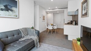 A modern living room with a grey sofa, a dining area with a white table and chairs, and a kitchen area with a stove and cabinets. - Dexter - Resort Living Gym, Sauna, Pool, Wi-Fi (Southbank)