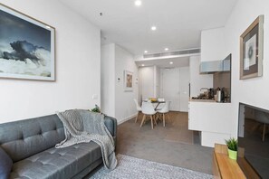 A modern living room with a grey sofa, a dining area with a white table and chairs, and a kitchen area with a stove and cabinets.