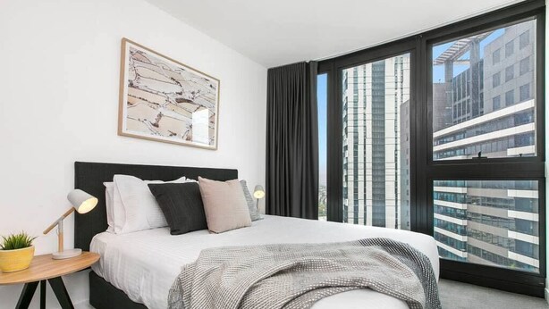 A modern hotel room with a large bed, a bedside table with a lamp, a framed artwork on the wall, and a view of the cityscape through large windows. - Dexter - Resort Living Gym, Sauna, Pool, Wi-Fi (Southbank)