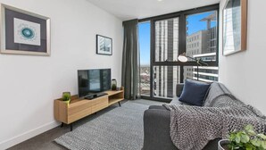 A modern living room with a flat-screen TV, a sofa, a coffee table, and a large window offering a city view. - Dexter - Resort Living Gym, Sauna, Pool, Wi-Fi (Southbank)