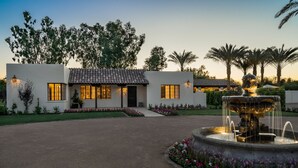 Una casa moderna con tejado de tejas, una fuente central y un jardín cuidado con diversas plantas y flores.