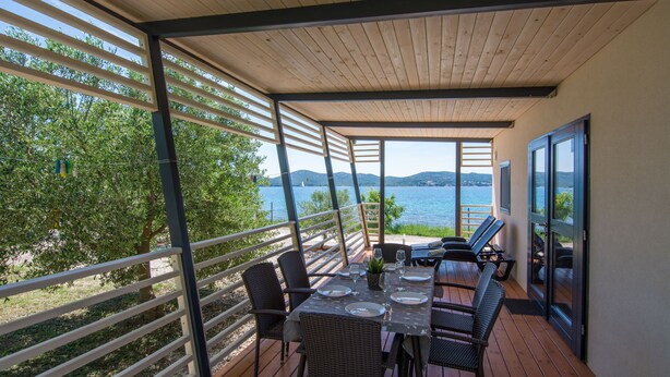 A covered outdoor dining area with a table set for two, overlooking a body of water and a mountainous landscape. - Mobile Home Plumeria 2 (Biograd)