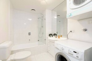 A bathroom with a toilet, bathtub, glass-enclosed shower, and a washing machine. - Madeline - Panoramic views * WiFi Gym Pool Parking (Southbank)
