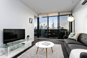 Een moderne woonkamer met een flatscreen televisie, een glazen salontafel, een grijze bank en een stadszicht door grote ramen.