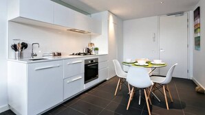 Moderni keittiö, jossa on valkoiset kaapit, ruokapöytä tuoleineen sekä pieni keittiösaareke, jossa on pesuallas ja liesi.