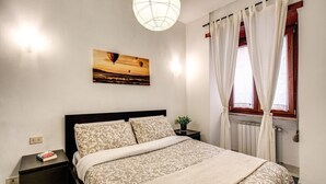 Un lit bien fait, recouvert d’une couvre-lit à motifs, une table de chevet avec un livre, un tableau représentant un coucher de soleil accroché au mur, et une fenêtre avec des rideaux.