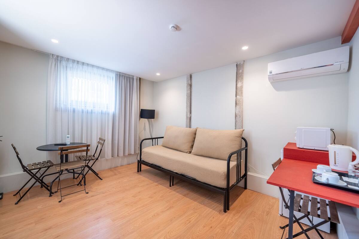 Un espacio habitable compacto con sofá, una mesa de comedor con sillas, una pequeña cocina con microondas y una ventana con cortinas.