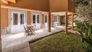 Un patio techado al aire libre con asientos, un pequeño jardín y un edificio con puertas blancas.