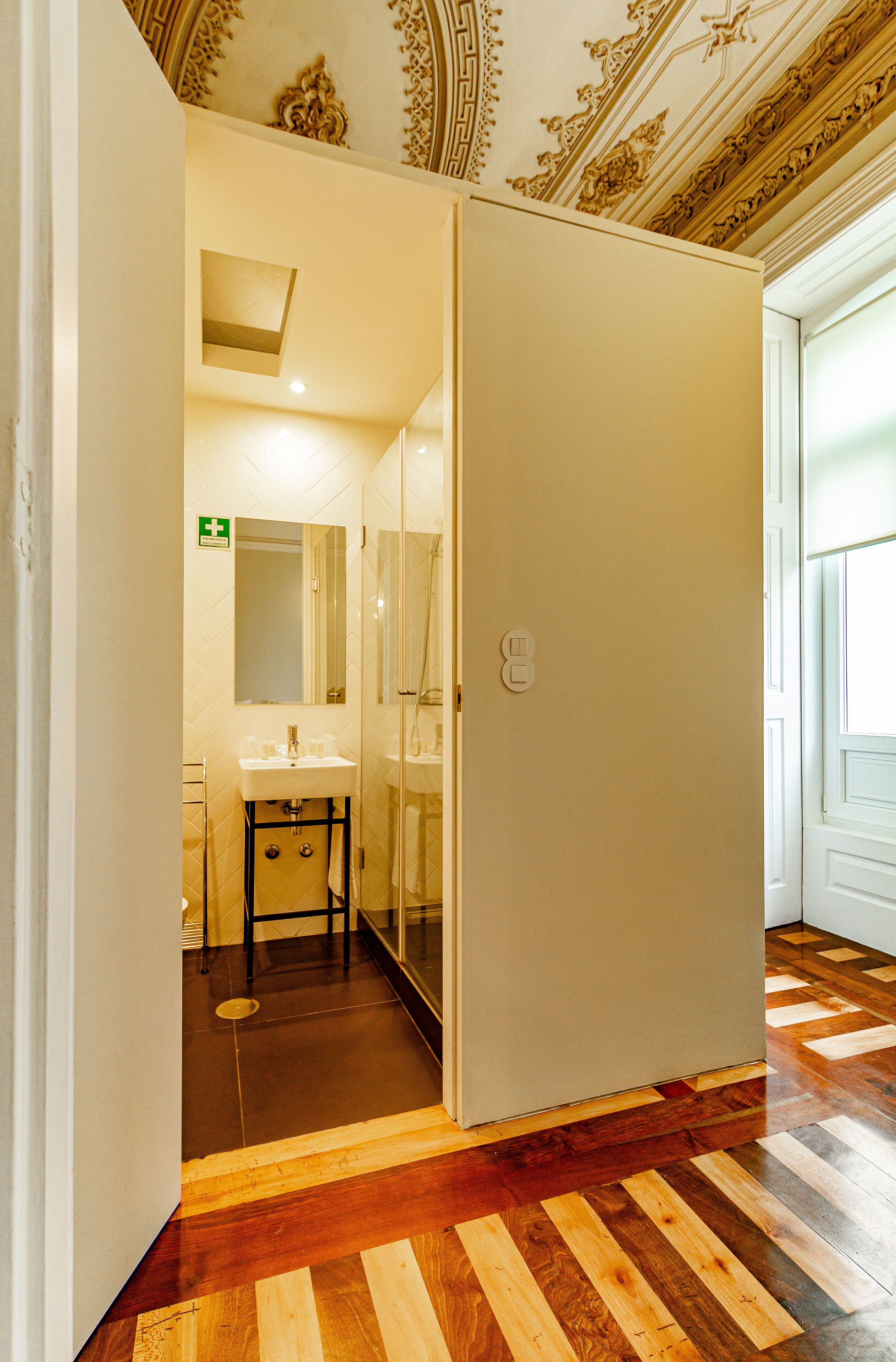 A hotel room with a wooden parquet floor, a white wall, a mirror, a sink, and a door leading to another room.