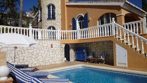 Un área de piscina con sillones de descanso y sombrillas, una torre de piedra y una escalera que lleva a un balcón.