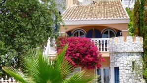Edificio de estilo mediterráneo con fachada de piedra, balcón con contraventanas azules y buganvilla rosa vibrante.