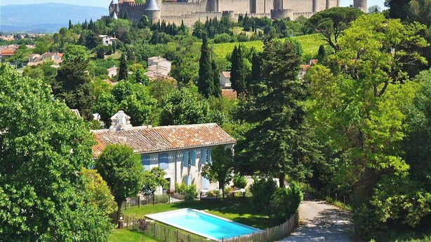 Un château entouré d’une végétation luxuriante, avec une piscine en premier plan.