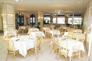 Un restaurante con mesas redondas, sillas de mimbre, ventanas amplias y candelabros.