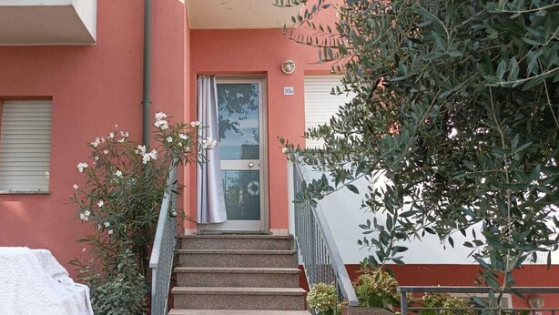 Um edifício vermelho com entrada por porta de vidro, escadaria e um carro coberto por uma lona branca.
