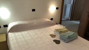 Uma cama bem arrumada com um cobre-leito estampado, uma pilha de toalhas dobradas e dois travesseiros em formato de coração sobre um chão de azulejos limpo.