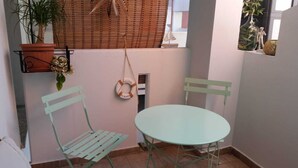 Uma pequena área externa com mesa redonda e duas cadeiras, uma planta em vaso e um objeto decorativo pendurado na parede.