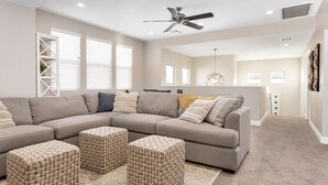 A living room with a sectional sofa, three woven ottomans, a ceiling fan, and a kitchen area in the background. - Desert Escape at Ladera (Washington)