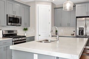 Una cocina moderna con armarios grises, electrodomésticos de acero inoxidable, encimera de mármol y una lámpara colgante.