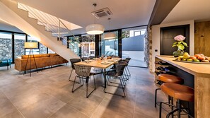 Uma área de refeições moderna com uma mesa grande, várias cadeiras, um sofá e um balcão bar com frutas e garrafas.