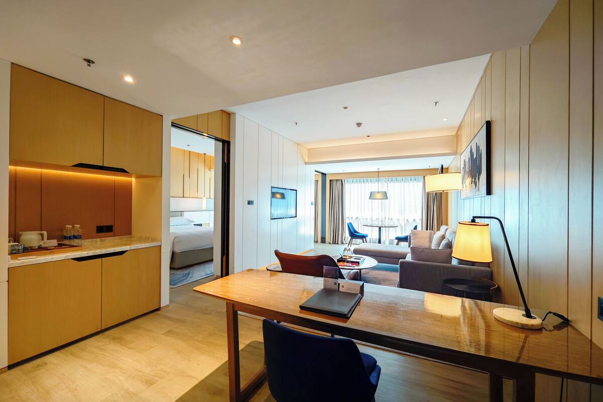 Een moderne hotelkamer met kitchenette, een eethoek, een zithoek met een bank en fauteuils, en een groot raam dat voor natuurlijk licht zorgt.