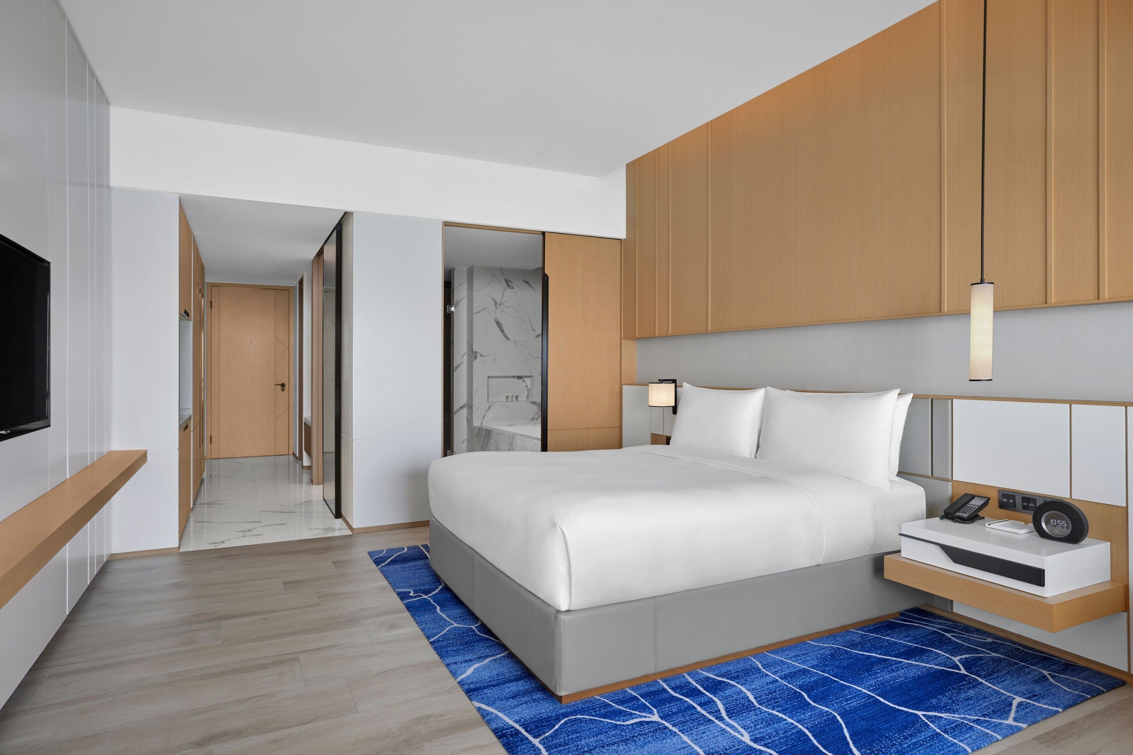 Ein modernes Hotelzimmer mit einem großen Bett, einem Fernseher, einem Nachttisch mit Telefon und Wecker und einem durchgehenden Türspalt sichtbaren Badezimmer.