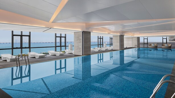 Een modern binnenzwembad met helderblauw water, witte ligbedden en grote ramen met uitzicht op zee.