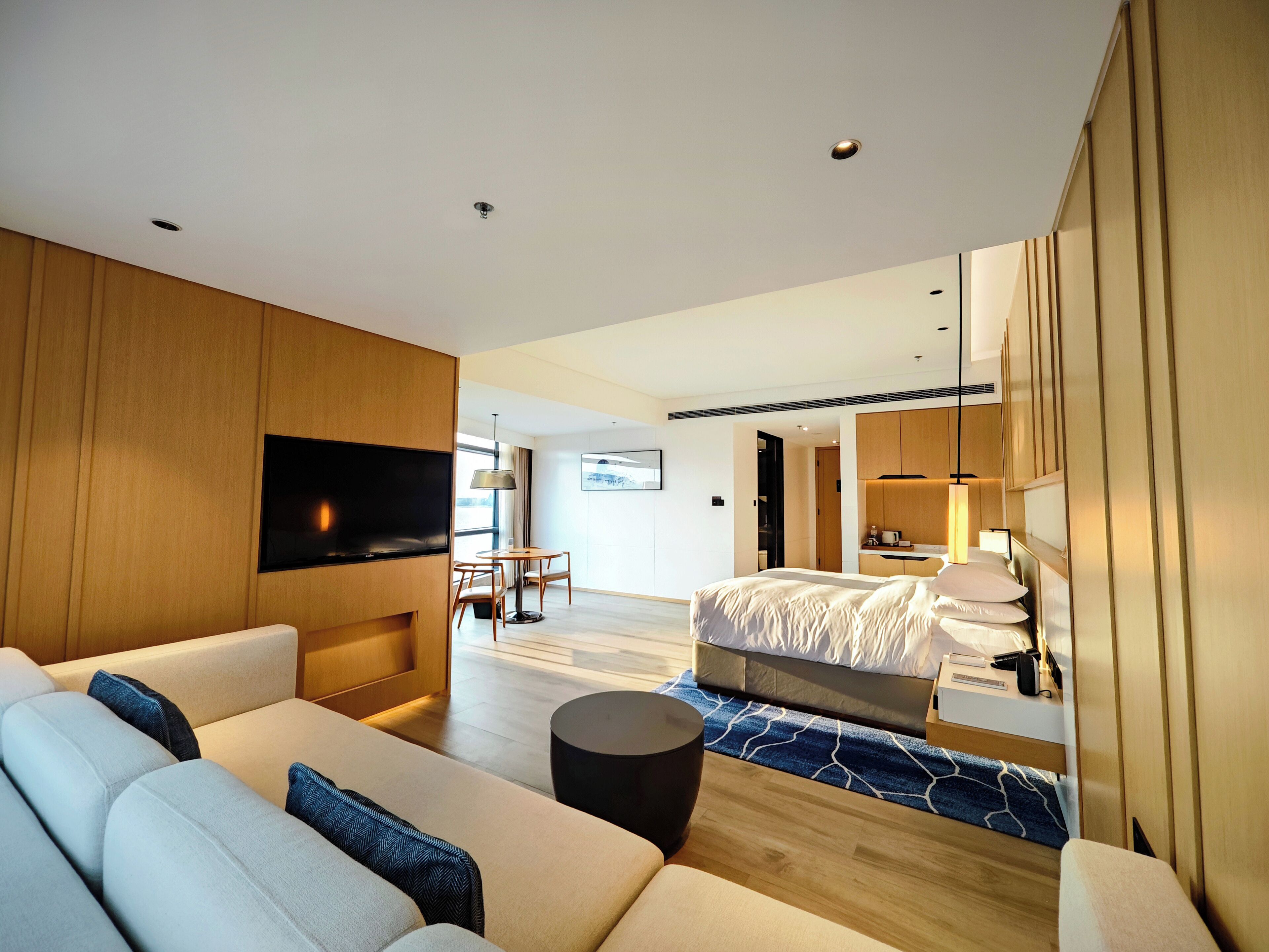 Una habitación de hotel moderna con sofá, cama, televisor montado en la pared y zona de comedor con mesa y sillas.