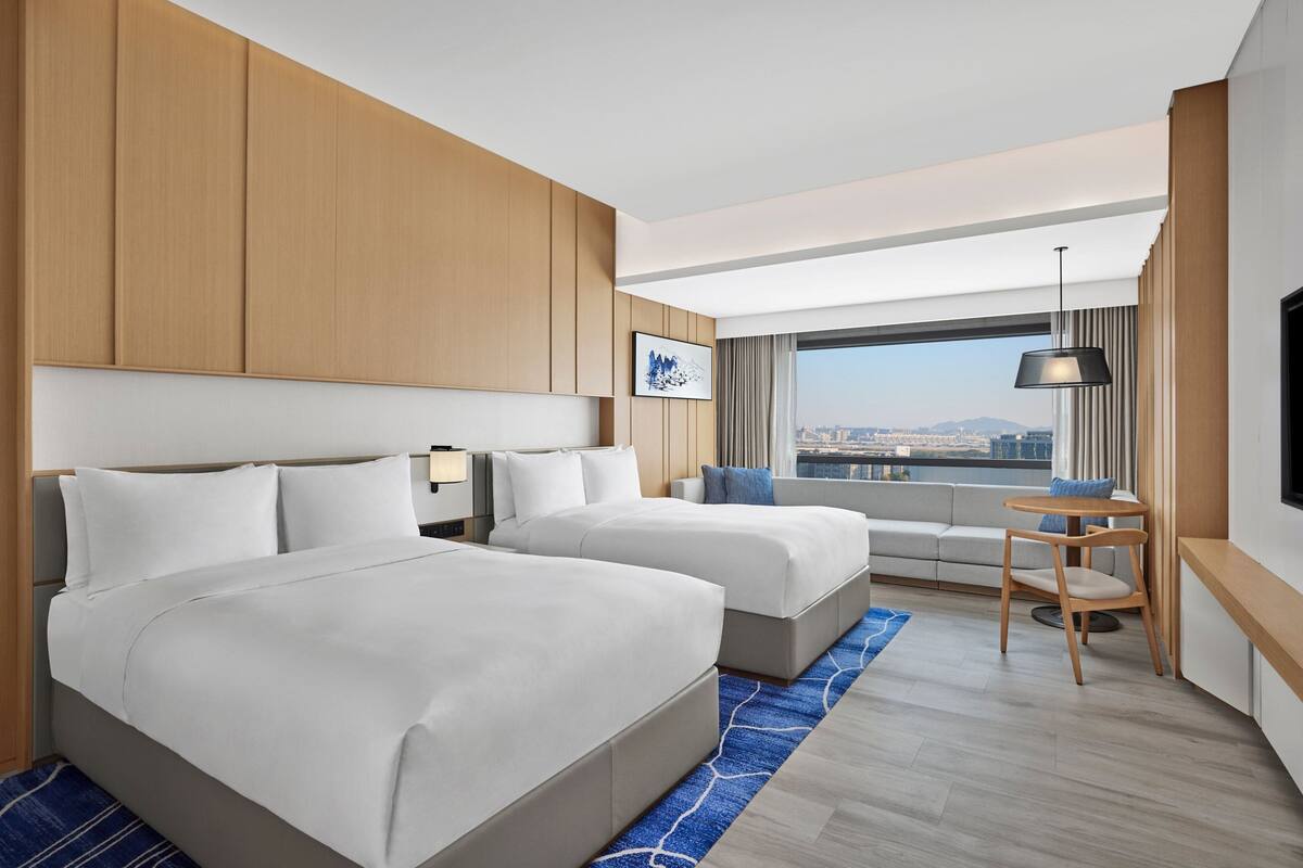 Een moderne hotelkamer met twee bedden, uitzicht op de stad, een klein tafeltje met een stoel en een groot raam met gordijnen.