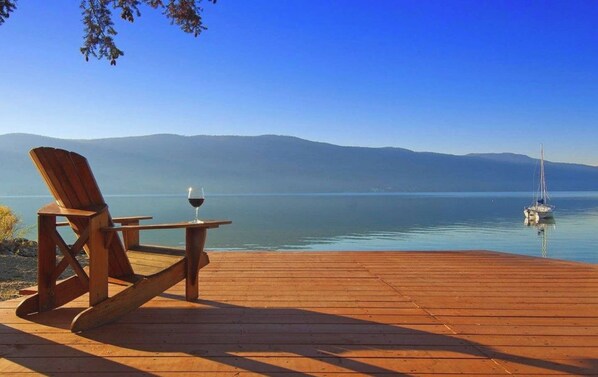 A wooden deck with a chair and a table, overlooking a calm lake with mountains in the background and a sailboat anchored near the shore.