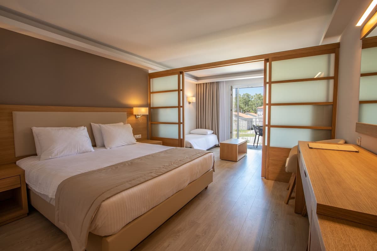 A hotel room with a large bed, a desk, and a chair. There is a window with curtains and a view of the outside.