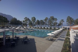 A pool area with lounge chairs and umbrellas, surrounded by trees and a mountain in the background. - Paleros Beach Resort (Aktio-Vonitsa)