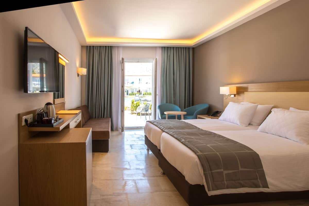 A hotel room with a bed, bedside tables, a desk, a television, and a balcony with chairs.