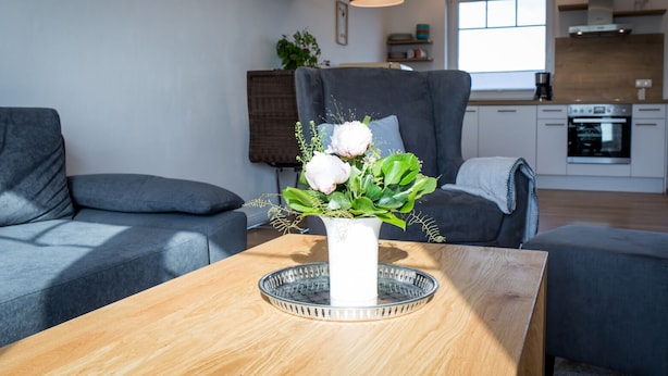 Ein modernes Wohnzimmer mit einer grauen Couch, einem Holztisch mit Blumenvase und einer Küche im Hintergrund.