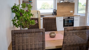 Ein Essbereich mit Korbstühlen, einem Tisch mit einer Vase und Blick auf eine Küche mit einer Kaffeemaschine.