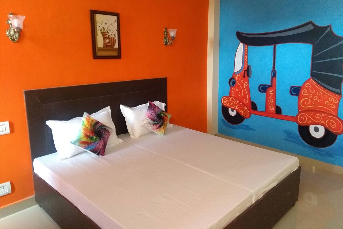 A hotel room with a bed, orange walls, a mural of a red scooter, and a framed picture on the wall.