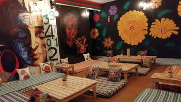 Una habitación con varias zonas de estar, incluyendo mesas y bancos, con llamativos murales florales y una gran pantalla con números.