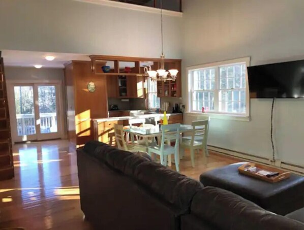 A living room with a dark brown sofa, a dining area with a white table and chairs, and a kitchen in the background. - Modern Country Cottage (West Kill)